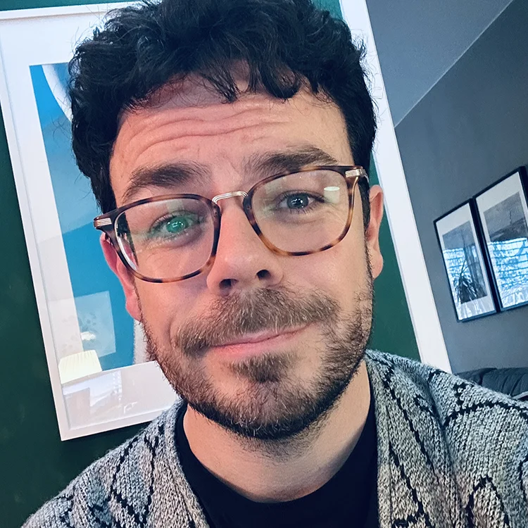 Nahaufnahme von Webdesigner Boris (IMPITAS) mit Brille und Bart vor einer markanten türkisen Wand. Das Selfie wirkt authentisch und nahbar, passend zu seiner Positionierung für sauberes Webdesign und technisches Handwerk ohne Schnickschnack.